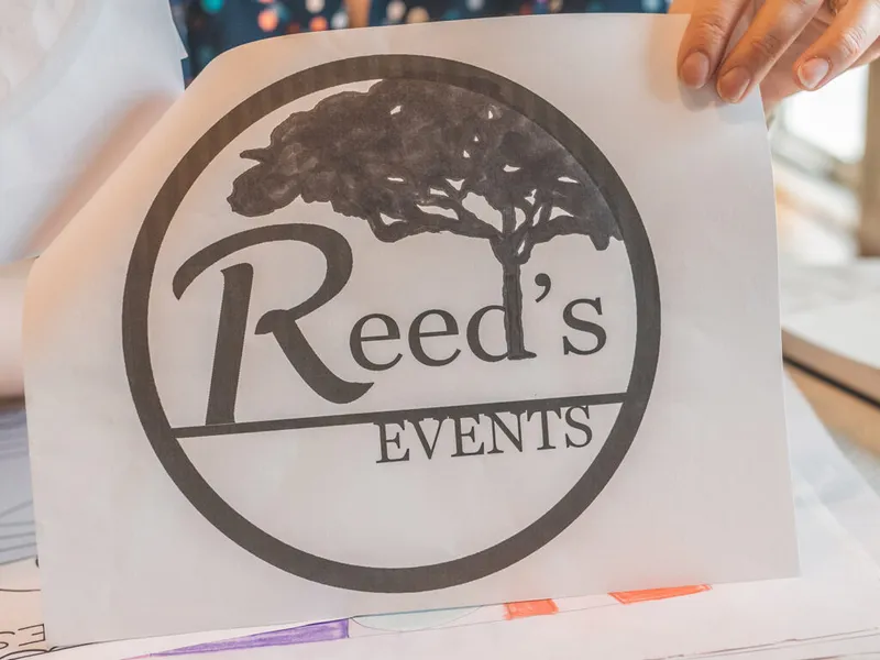 Hand holding a paper print of the Reed's Events circular logo with a tree illustration