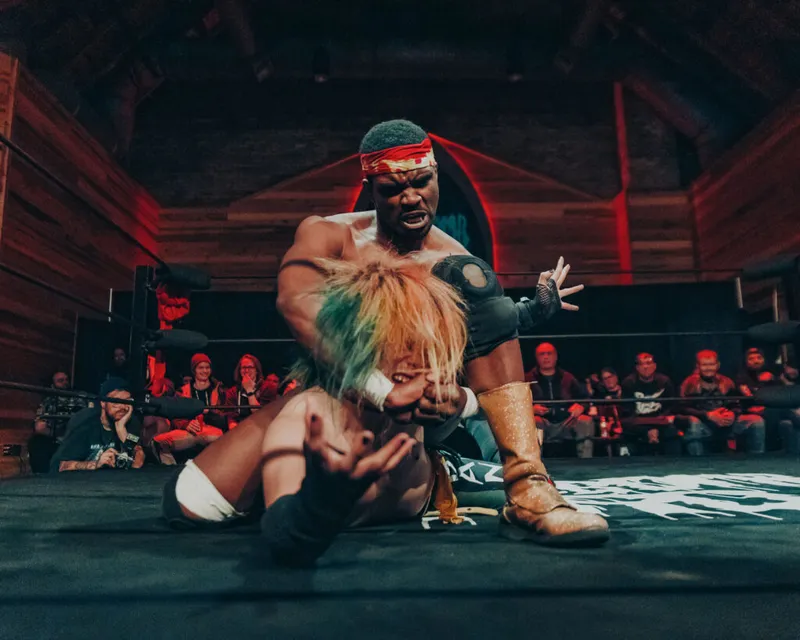 Pro wrestler in red headband pins opponent with multicolored hair on mat before a ringside crowd