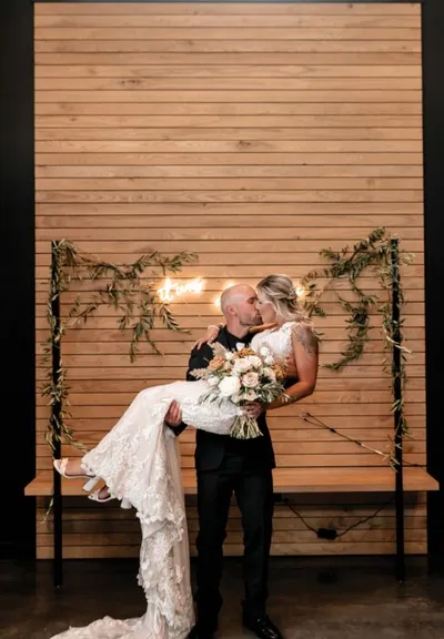 Groom in black suit lifting bride in lace gown as they kiss before a wood slat wall with greenery