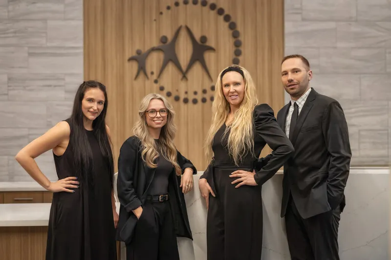 Four professionals in black attire posed in a modern office lobby with a people-figure logo on the wall