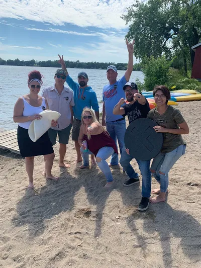 Group of seven adults posing on a sandy beach beside a lake with kayaks behind them