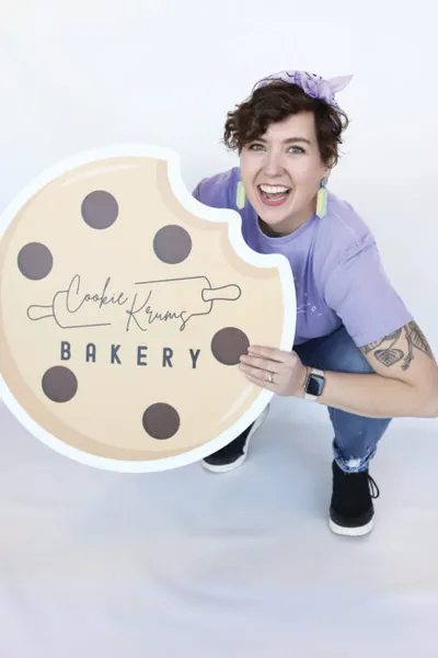 Smiling woman in purple tee crouching behind a large Cookie Krums Bakery sign