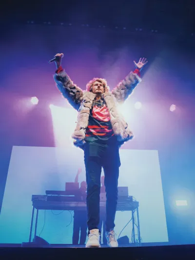 Male performer in fur coat raises both arms on a fog-lit concert stage holding a microphone