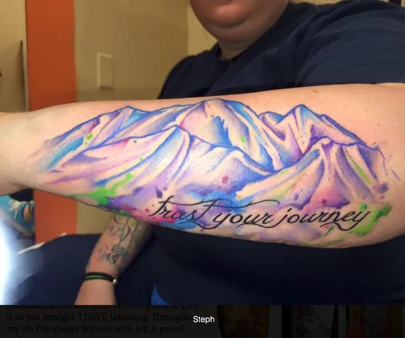 Forearm tattoo of watercolor mountains with cursive text reading trust your journey