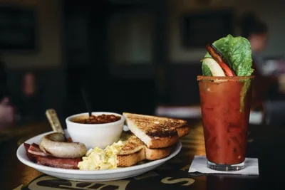 Bloody Mary garnished with bacon and lettuce beside a brunch plate with toast, sausage, and a small bowl of chili