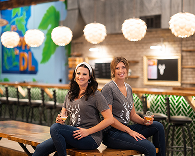 Two women in matching gray deer-logo tees holding beers in a brewery taproom