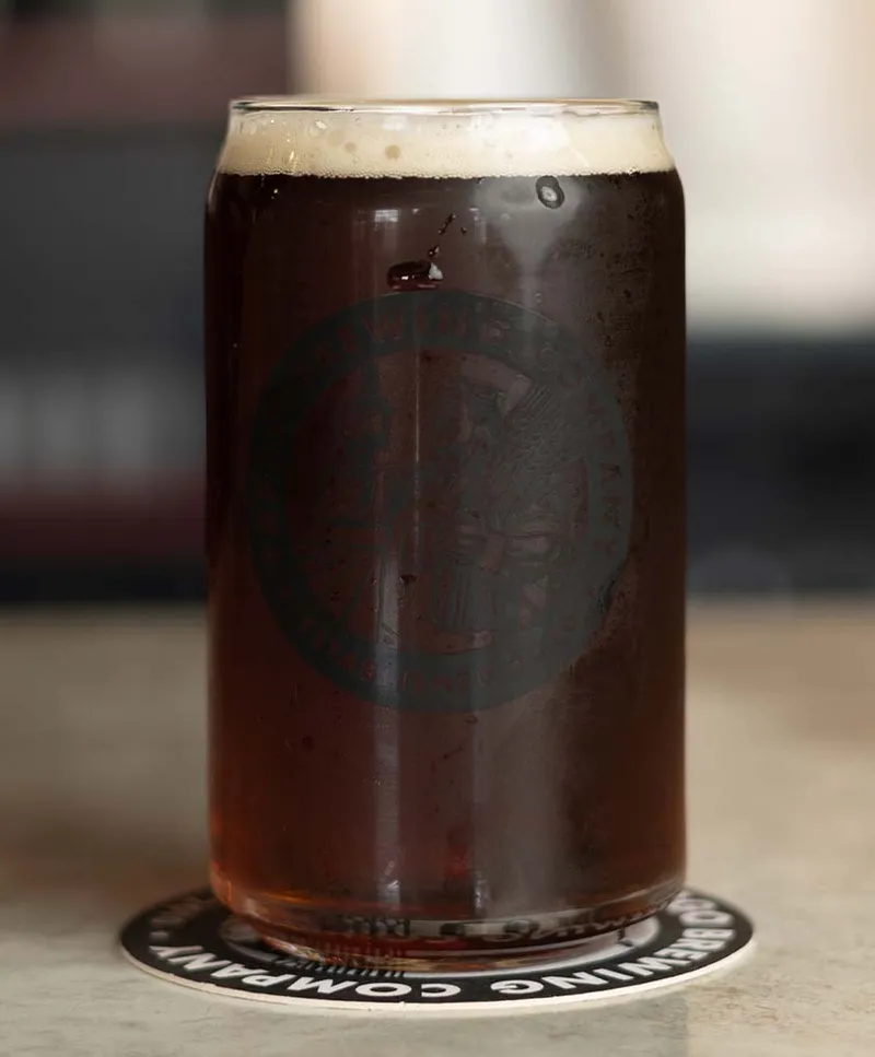 Dark beer with a thin foam head in a branded glass pint cup on a brewery coaster
