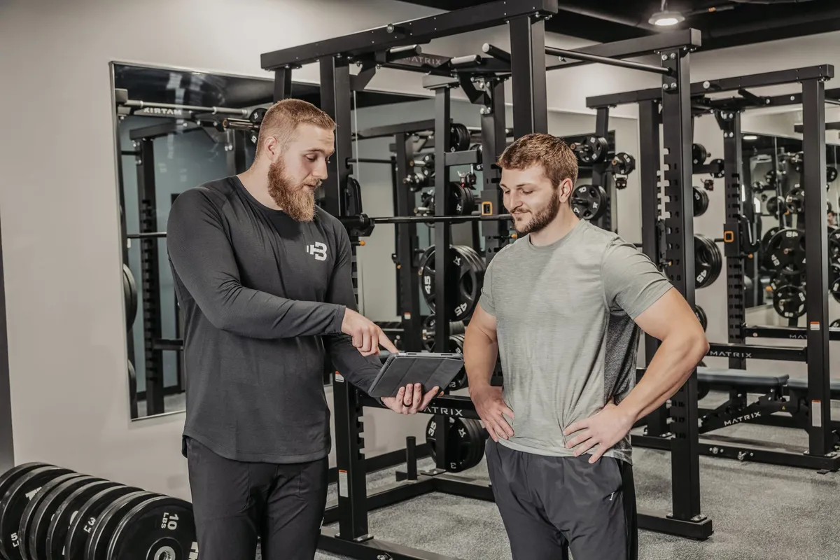 A trainer points to a tablet while consulting with a client in a well-equipped weight room