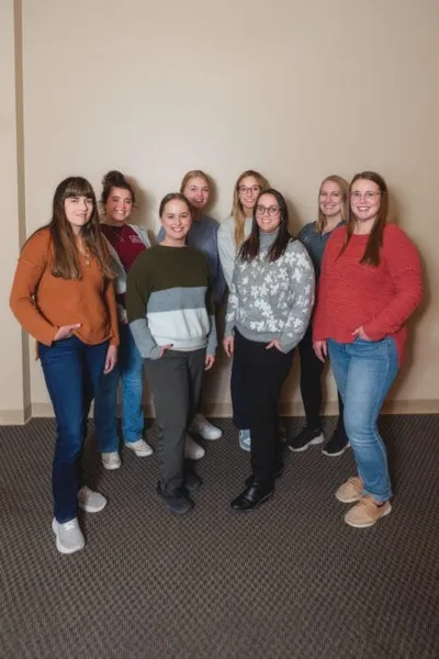 Eight young women posing together in two rows against a beige wall