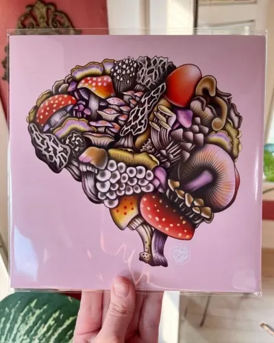 Hand holding a square art print of colorful mushrooms arranged in a brain shape on a pink background