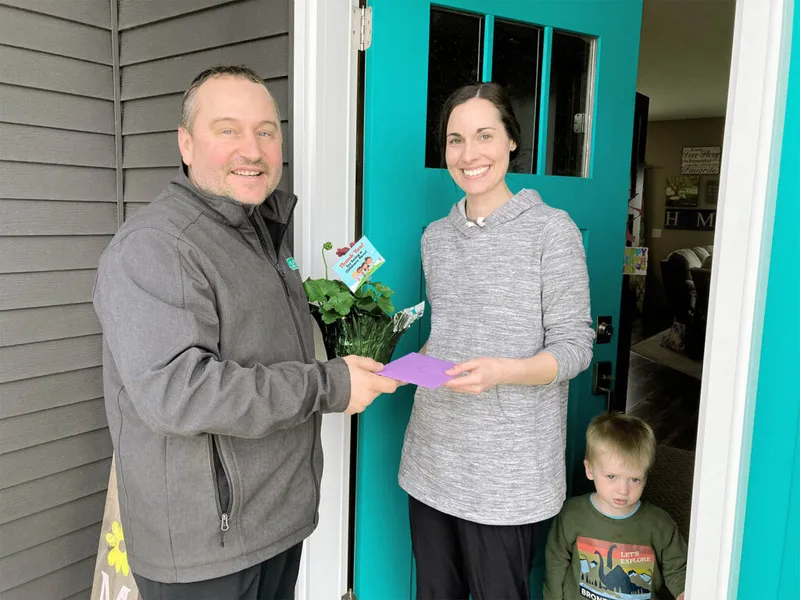 Man hands a small potted plant and purple envelope to a smiling woman at a teal front door, young boy beside her