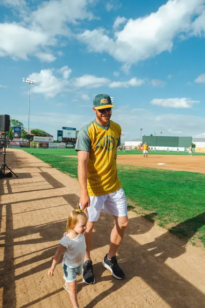 Man in yellow pinstripe baseball jersey holds a toddler's hand on a softball field