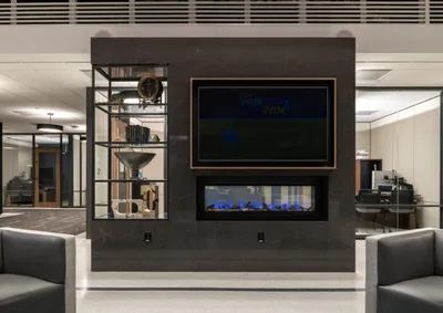 Dark stone accent wall with wall-mounted TV, electric fireplace, open shelving, and gray lounge chairs