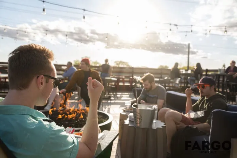 People socializing on an outdoor patio around a fire pit under string lights at sunset
