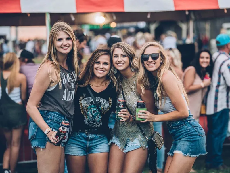 Four smiling women in denim shorts pose together holding canned drinks at an outdoor music festival