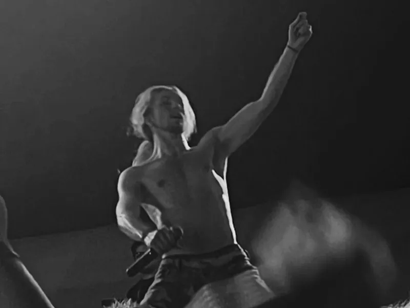 Shirtless performer with long blond hair raises one arm on a darkened stage