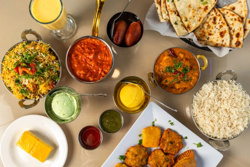 Overhead spread of Indian dishes including curry, biryani, naan, fried appetizers, chutneys, and a mango lassi