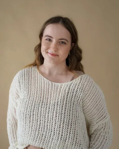 Young woman with wavy brown hair smiling, wearing a cream knit sweater against a beige background
