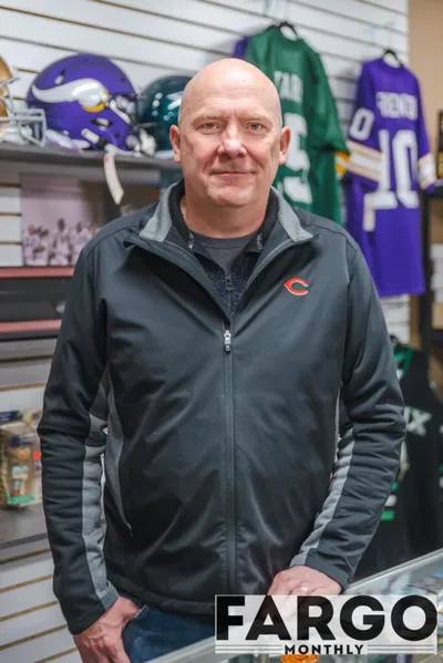 Bald man in a Chicago Bears jacket standing in a sports memorabilia shop with jerseys and helmets behind him