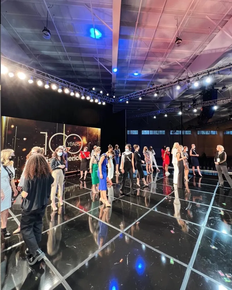 Masked pageant contestants lined up on a reflective stage beneath a '100 Years Miss America' backdrop