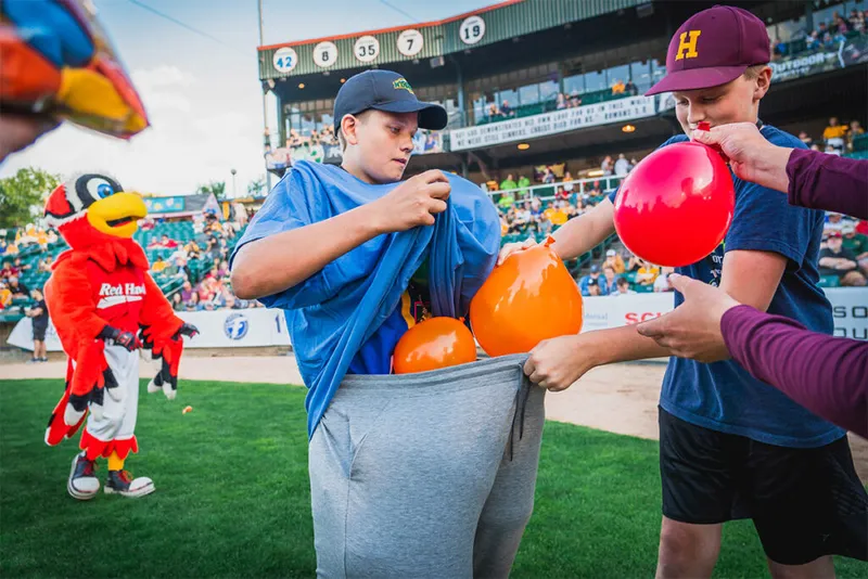 Two boys stuff orange balloons into oversized sweatpants during a field game, with a red bird mascot visible behind them