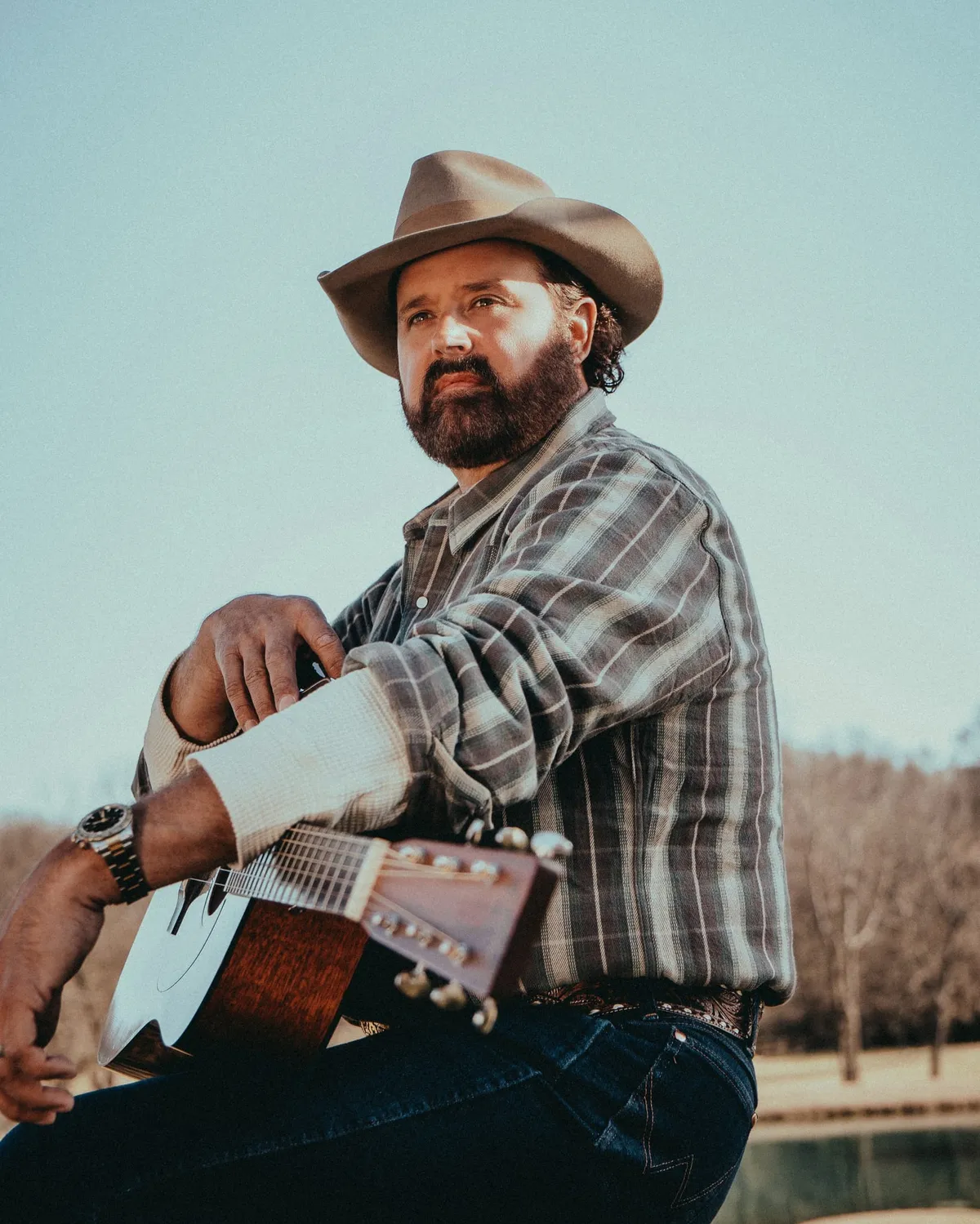 Bearded man in a tan cowboy hat and plaid shirt holding an acoustic guitar outdoors