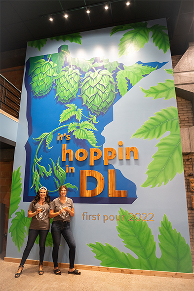 Two women holding drinks stand before a large mural reading 'it's hoppin in DL, first pour 2022
