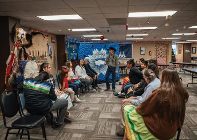 A speaker addresses a circle of seated attendees in a community room with Indigenous cultural items on display