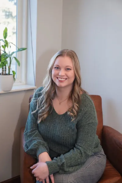 Smiling young woman with long blonde hair seated in a brown leather chair wearing a teal sweater