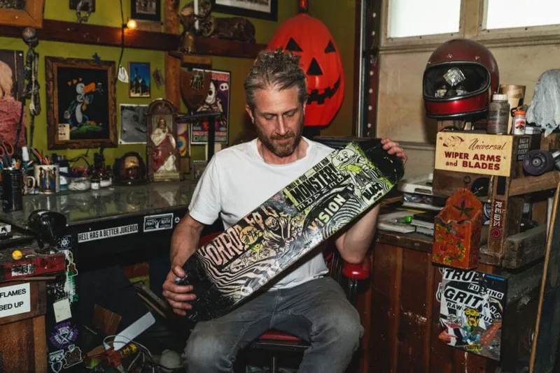 Man in white t-shirt examining a graphic-covered skateboard deck in a cluttered workshop