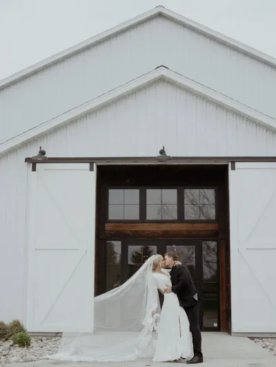 Couple kissing in wedding attire before a white barn venue with sliding doors