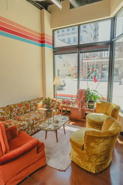 Retro lounge with floral sofa, velvet chairs, and rainbow wall stripes beside large street-facing windows