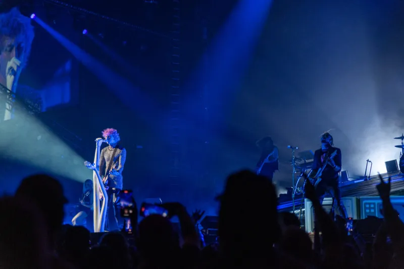 Singer with pink hair performs at a microphone under blue stage lights before a packed crowd