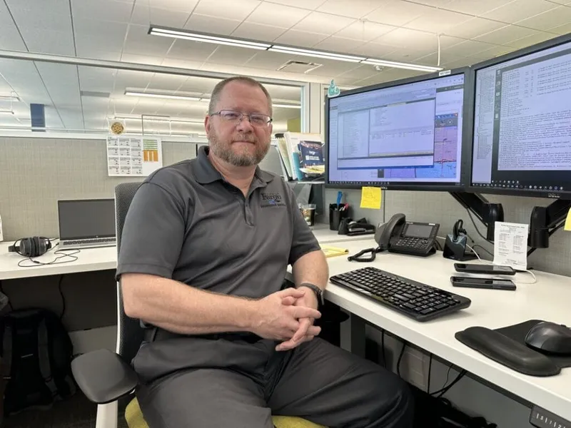 Man in gray polo shirt seated at an office cubicle desk with dual monitors