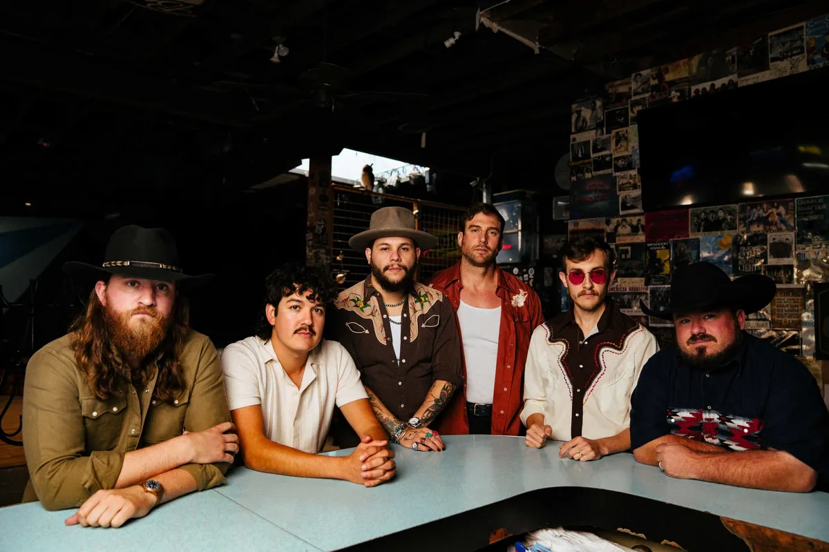 Five men in western-style shirts and cowboy hats seated around a table in a dimly lit venue with flyers covering the walls