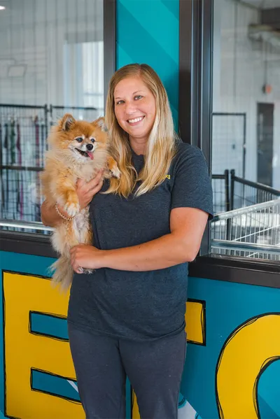Smiling blond woman holding a fluffy tan Pomeranian inside a bright teal and yellow pet facility