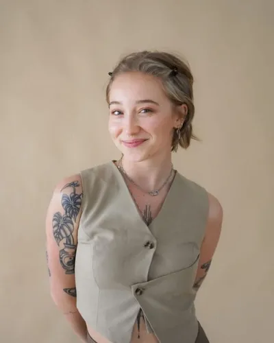 Smiling woman with tattooed arms in a sage sleeveless wrap top against a neutral background