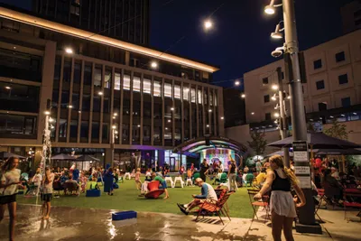 Crowd gathered on a green urban plaza at night beneath string lights and tall buildings