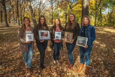 Five women holding framed photos stand among autumn trees covered in fallen leaves