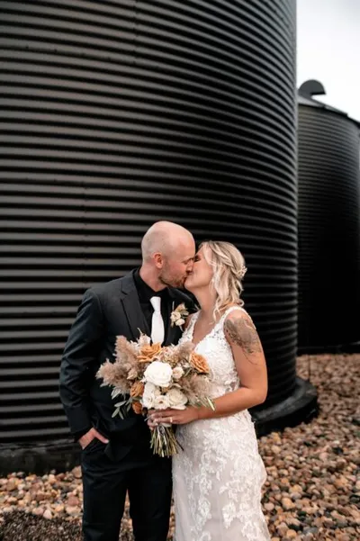 Bride and groom kissing in front of dark grain silos, she holds a boho bouquet of pampas grass and white roses