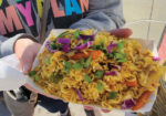 Hands holding a large colorful noodle salad topped with fresh herbs and edible flowers
