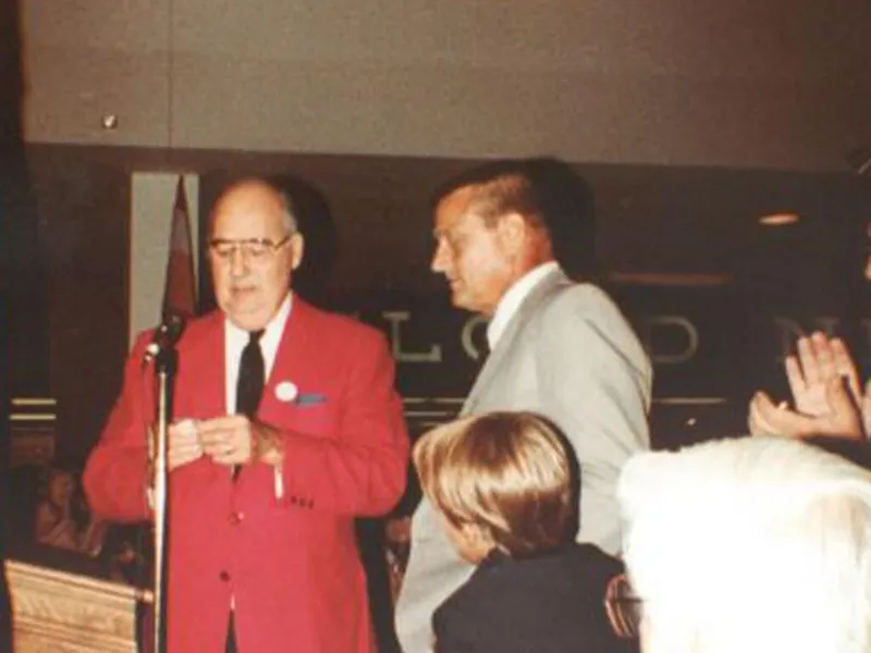Man in red blazer speaking at a microphone beside another man in a gray suit at an indoor event