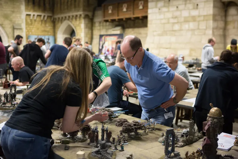 Two people lean over a table of miniature wargaming figures inside a stone-walled hall