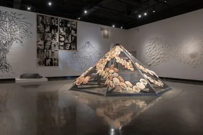 Gallery installation with a pyramid sculpture covered in shells, flanked by large ink drawings on white walls
