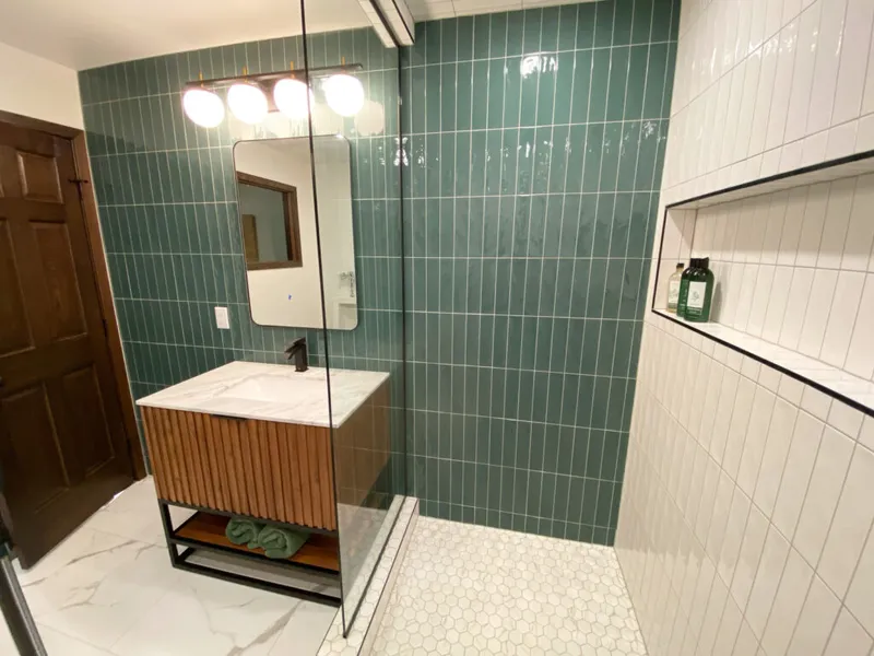 Renovated bathroom with teal vertical wall tiles, wood-front vanity with marble top, and glass shower enclosure