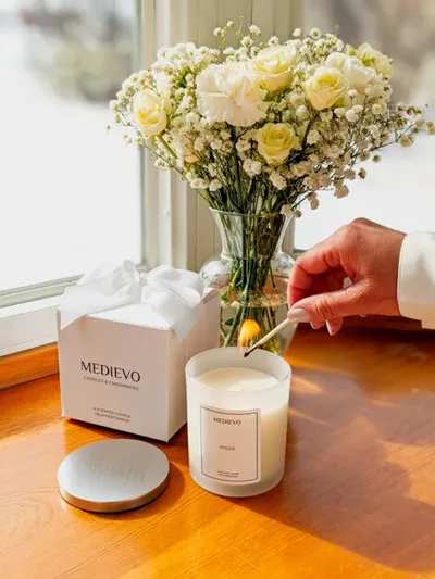 Hand lighting a Medievo candle with a match beside white roses and a gift box