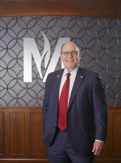 Man in navy suit and red tie smiling before a large decorative M logo on a textured wall