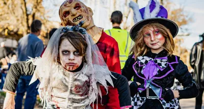 Two children in zombie and top-hat Halloween costumes pose with a tall masked figure outdoors