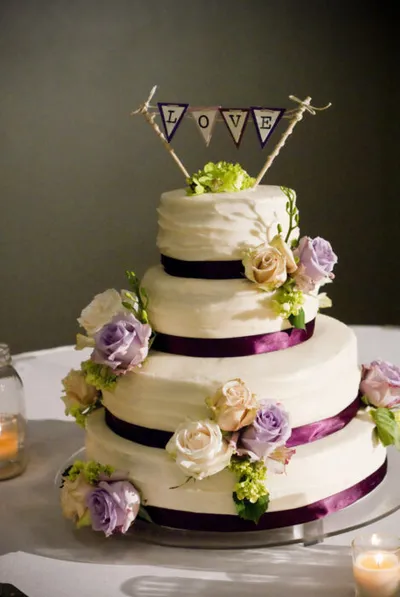 Three-tier white wedding cake with purple ribbons, lavender roses, and a "LOVE" bunting topper