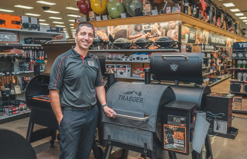 Smiling store employee in dark polo stands beside a Traeger pellet grill inside a retail store